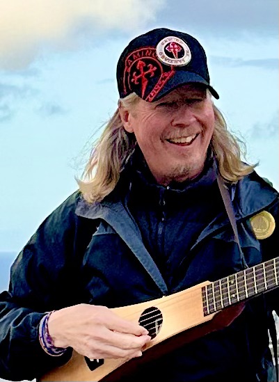 Dave Hall smiling in a Camino cap, playing a travel guitar by the sea